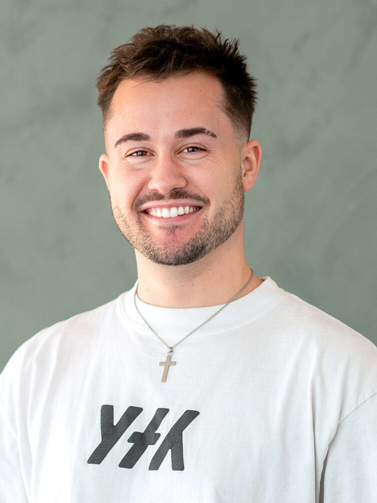 Thomas Russ' headshot. He wears a white t-shirt and cross necklace. 
