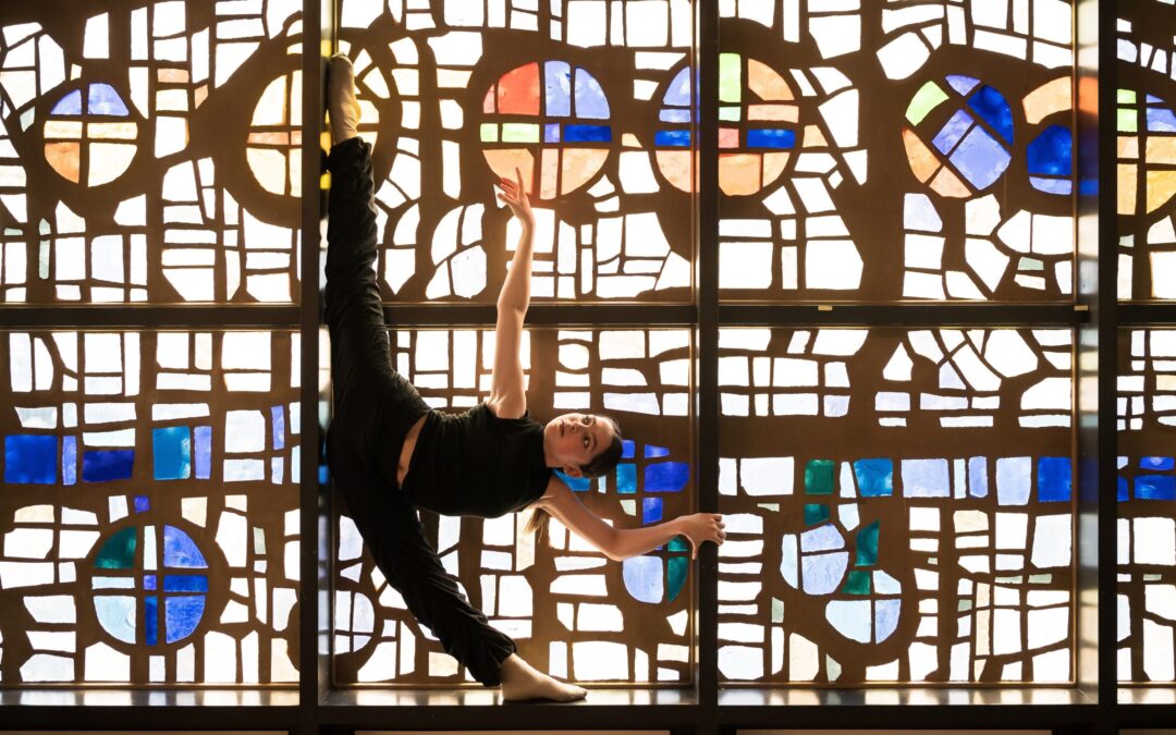 In front of a large stained glass window, a dancer poses in a standing oversplit with one arm lifted up as she holds onto one of the window's mullion with the other. She wears all black.