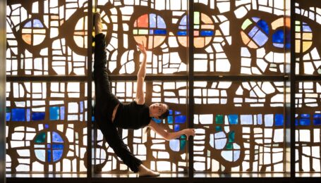 In front of a large stained glass window, a dancer poses in a standing oversplit with one arm lifted up as she holds onto one of the window's mullion with the other. She wears all black.
