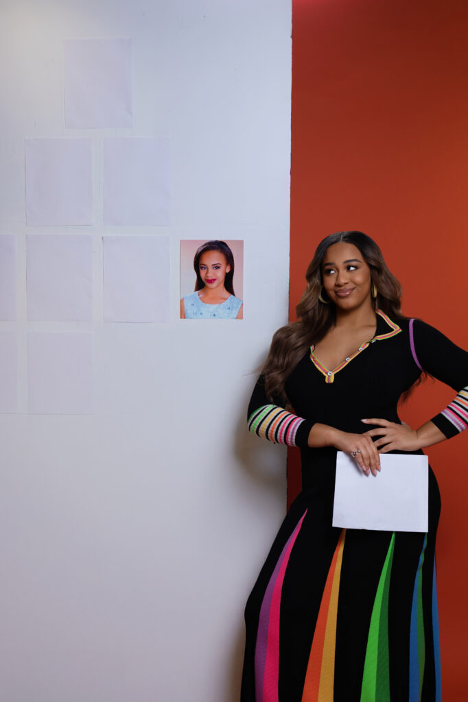 Nia Sioux, in a long black dress with colorful panels in the skirt and rings at the lower sleeve, stands with one hand on her hip, the other holding a piece of paper. On the wall behind her is a pyramid of headshots with paper over top of them, except for the one at bottom right, which is Sioux's own childhood headshot.