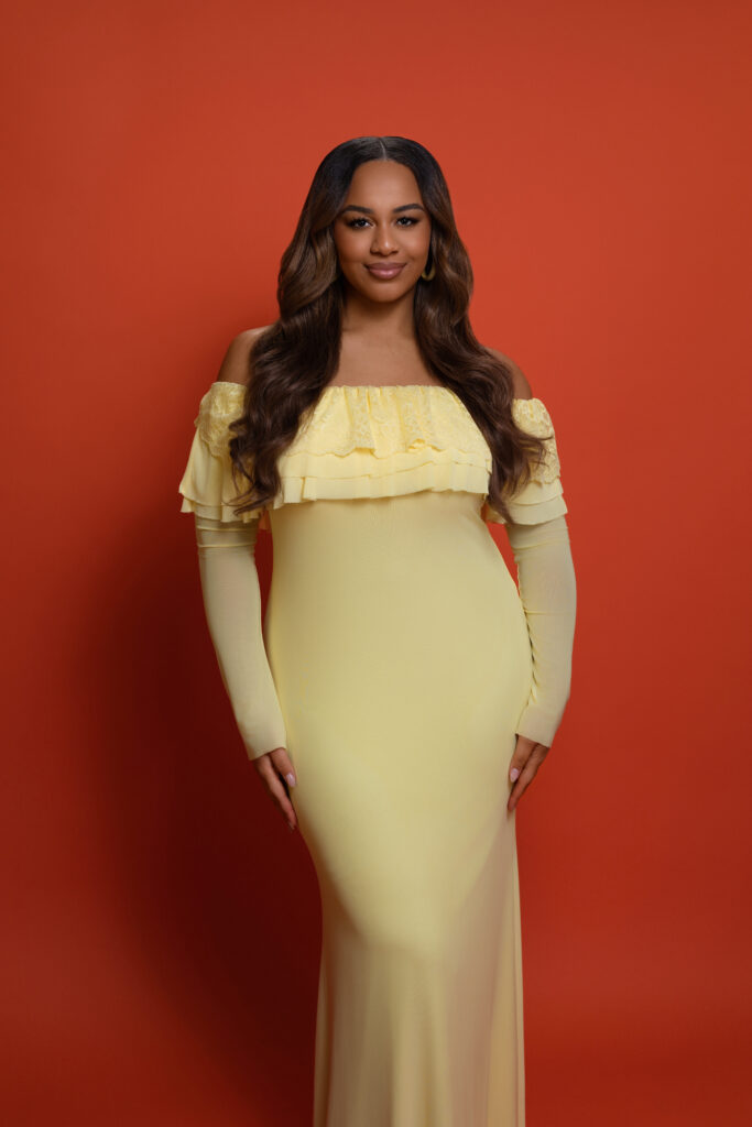 In front of a bright red background, Nia Sioux poses in a light yellow dress with one knee casually popped, arms at her sides.