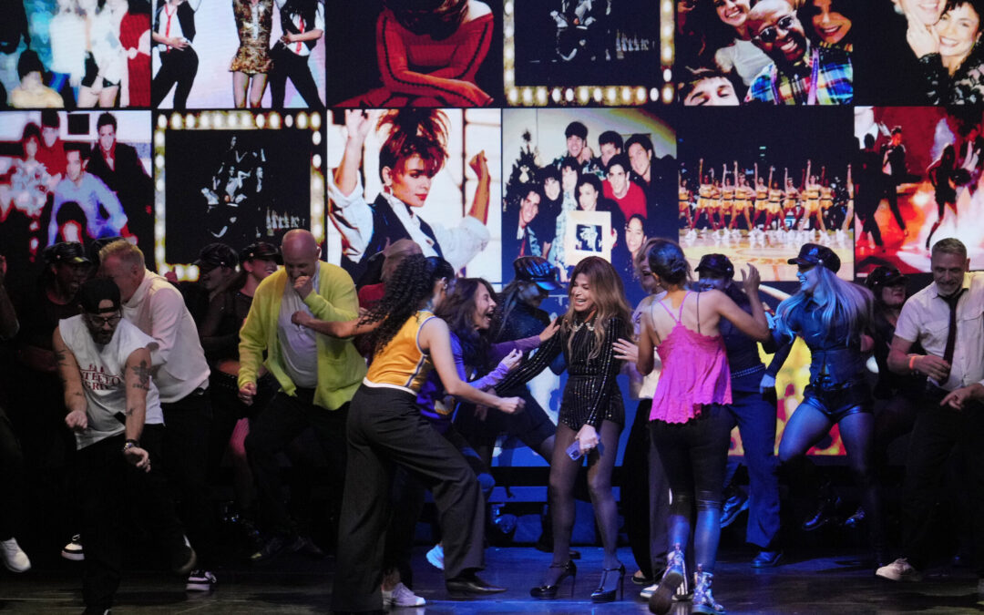 Onstage, a group of dancers move and celebrate together, congratulating Paula Abdul, who laughs and stands in the middle of the stage. Behind them is a large projection of throwback photos of Abdul throughout the years.