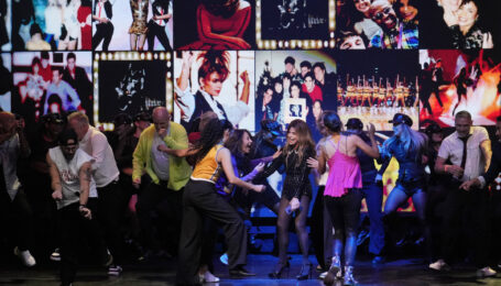 Onstage, a group of dancers move and celebrate together, congratulating Paula Abdul, who laughs and stands in the middle of the stage. Behind them is a large projection of throwback photos of Abdul throughout the years.