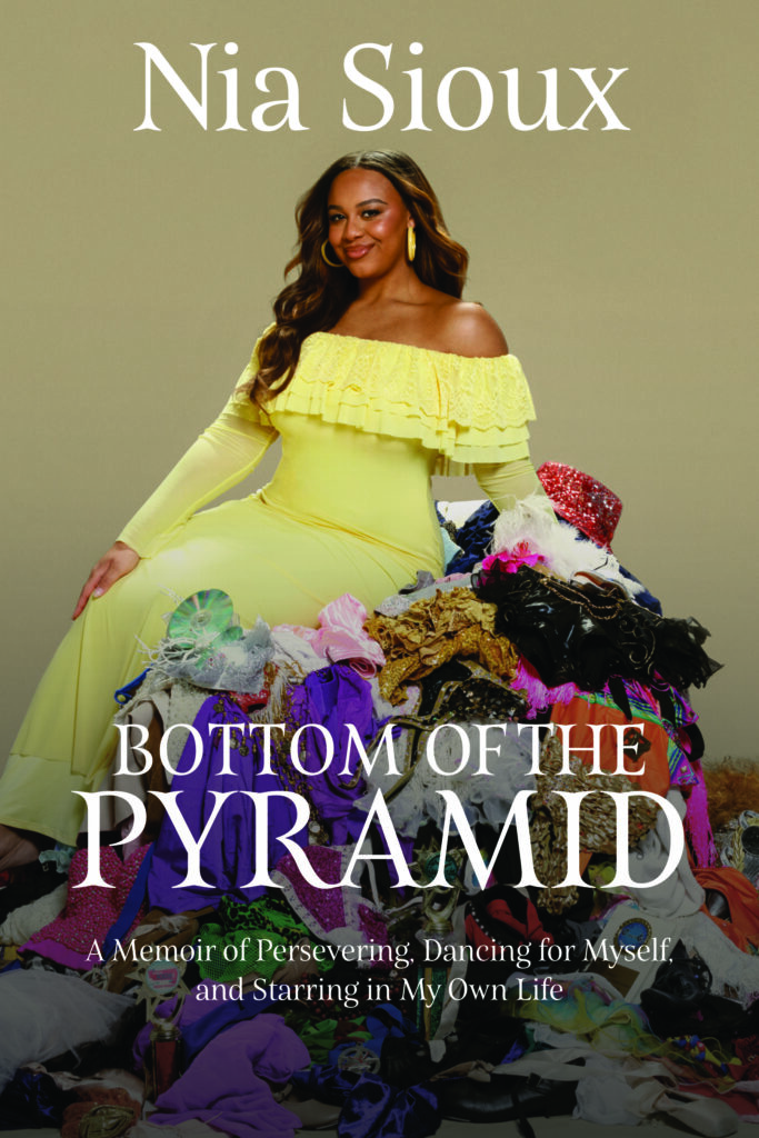 The cover of Nia Sioux's memoir, "Bottom of the Pyramid: A Memoir of Persevering, Dancing for Myself, and Starring in My Own Life." Sioux, in a light yellow dress, sits on the side of her hip atop a large pile of old children's dance costumes. She smiles at the camera.