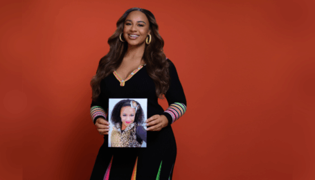 In front of a red background, Nia Sioux poses holding her childhood headshot. She wears a long black dress with colorful pleats in the skirt and rings on the lower sleeve.