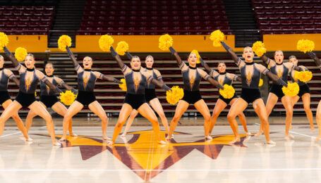 On a basketball court, the ASU dance team performs a pom routine wearing dark red leotards with gold and white sparkles. They dance with yellow poms and bend their knees with one arm up and the other extended down on the diagonal.