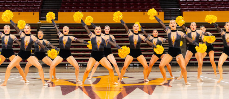 On a basketball court, the ASU dance team performs a pom routine wearing dark red leotards with gold and white sparkles. They dance with yellow poms and bend their knees with one arm up and the other extended down on the diagonal.