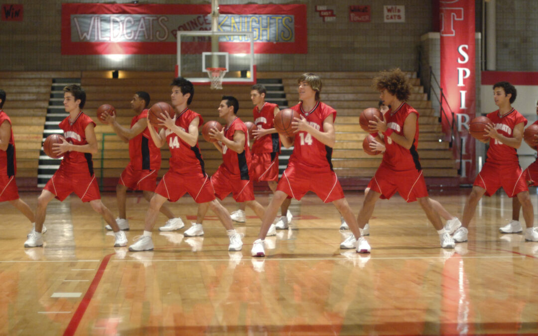 On a high school basketball court, a group of dancers in red basketball uniforms dance together. They spread their legs wide and flip basketballs over between their hands.