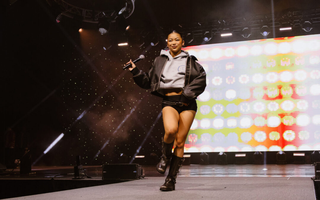 Kira Chan struts down a runway at a dance convention, holding a microphone in one hand. She wears combat boots, black shorts with a belt, and a gray sweatshirt under a leather jacket.