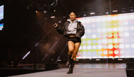 Kira Chan struts down a runway at a dance convention, holding a microphone in one hand. She wears combat boots, black shorts with a belt, and a gray sweatshirt under a leather jacket.
