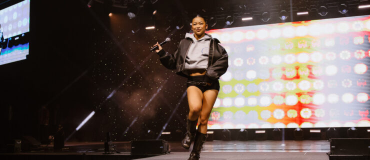 Kira Chan struts down a runway at a dance convention, holding a microphone in one hand. She wears combat boots, black shorts with a belt, and a gray sweatshirt under a leather jacket.