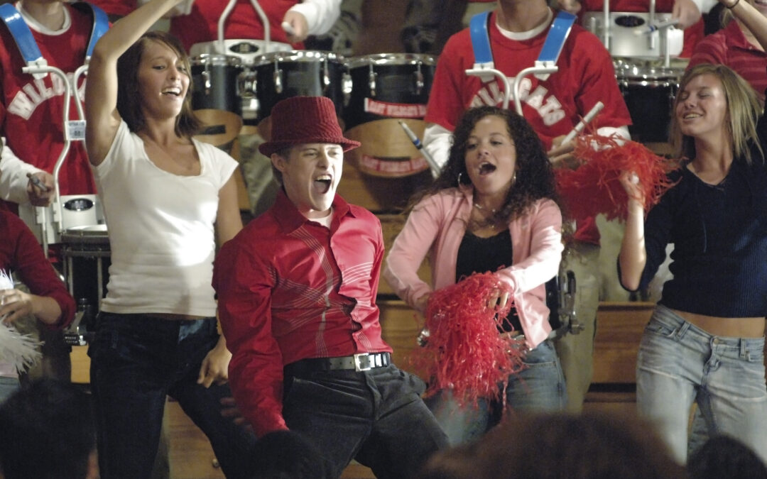 A still from "High School Musical" (which celebrates its 20th anniversary this year) showing a large group of students celebrating in a gym with pom-pons and streamers. Lucas Grabeel, in a red button down, a matching fedora, and black pants, pumps his arms down and opens his mouth as he sings.