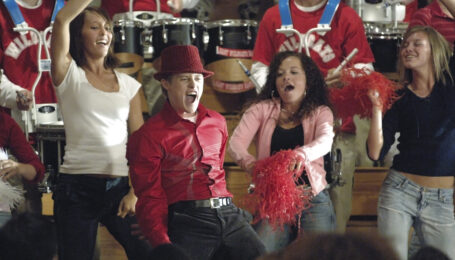 A still from "High School Musical" (which celebrates its 20th anniversary this year) showing a large group of students celebrating in a gym with pom-pons and streamers. Lucas Grabeel, in a red button down, a matching fedora, and black pants, pumps his arms down and opens his mouth as he sings.