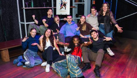 Ten dancers wearing street clothes huddle together for a group photo, making funny faces and laughing.