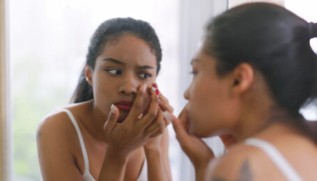 A close-up shot of a young woman looking at her reflection in the mirror. She is upset because she has a pimple.