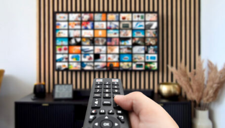 A close-up shot of someone's hand holding a TV remote, pointed toward the screen on the opposite wall.