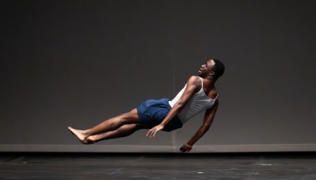 Nnamdi Nwagwu weaing a white vest and blue shorts suspended in a sideways falling pose on stage.