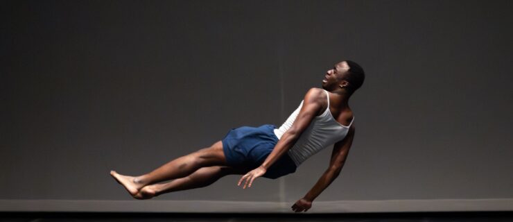 Nnamdi Nwagwu weaing a white vest and blue shorts suspended in a sideways falling pose on stage.