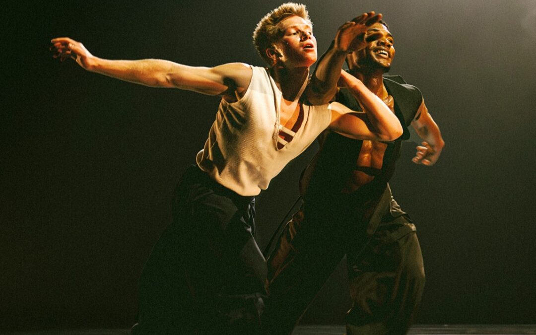 Joziah German and Sidney Chuckas performing onstage. They lunge forward with German holding onto Chuckas' arm. The two dancers wear black pants and white or black tanks.