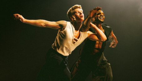 Joziah German and Sidney Chuckas performing onstage. They lunge forward with German holding onto Chuckas' arm. The two dancers wear black pants and white or black tanks.