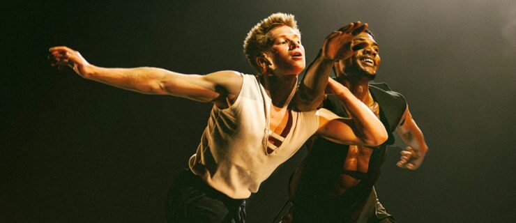 Joziah German and Sidney Chuckas performing onstage. They lunge forward with German holding onto Chuckas' arm. The two dancers wear black pants and white or black tanks.