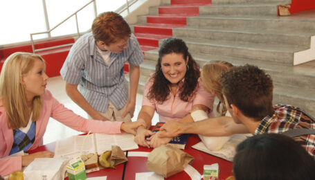 A still from "High School Musical" shows six teenagers sitting at a round lunch table in a school cafeteria. The students crowd around one, who smiles as she addresses them, and they reach out their arms to rest one hand on hers.
