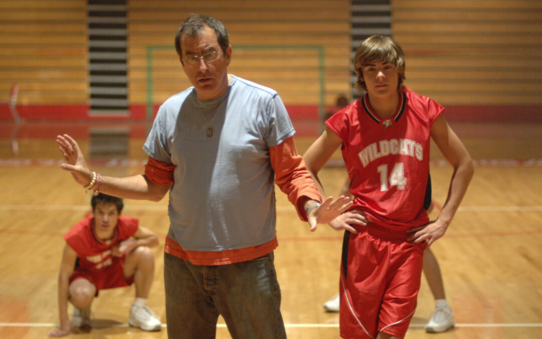 In a high school basketball gym, Kenny Ortega stands with his arms gently posed at either side. He looks slightly down and speaks to three dancers, clad in red and white basketball uniforms, who stand or crouch behind him.