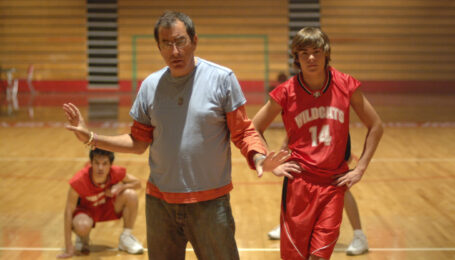 In a high school basketball gym, Kenny Ortega stands with his arms gently posed at either side. He looks slightly down and speaks to three dancers, clad in red and white basketball uniforms, who stand or crouch behind him.