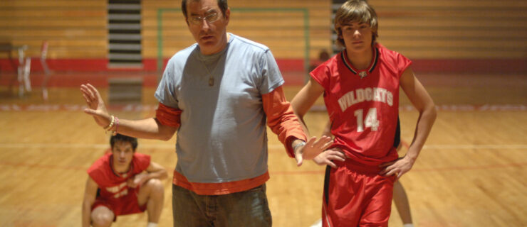 In a high school basketball gym, Kenny Ortega stands with his arms gently posed at either side. He looks slightly down and speaks to three dancers, clad in red and white basketball uniforms, who stand or crouch behind him.
