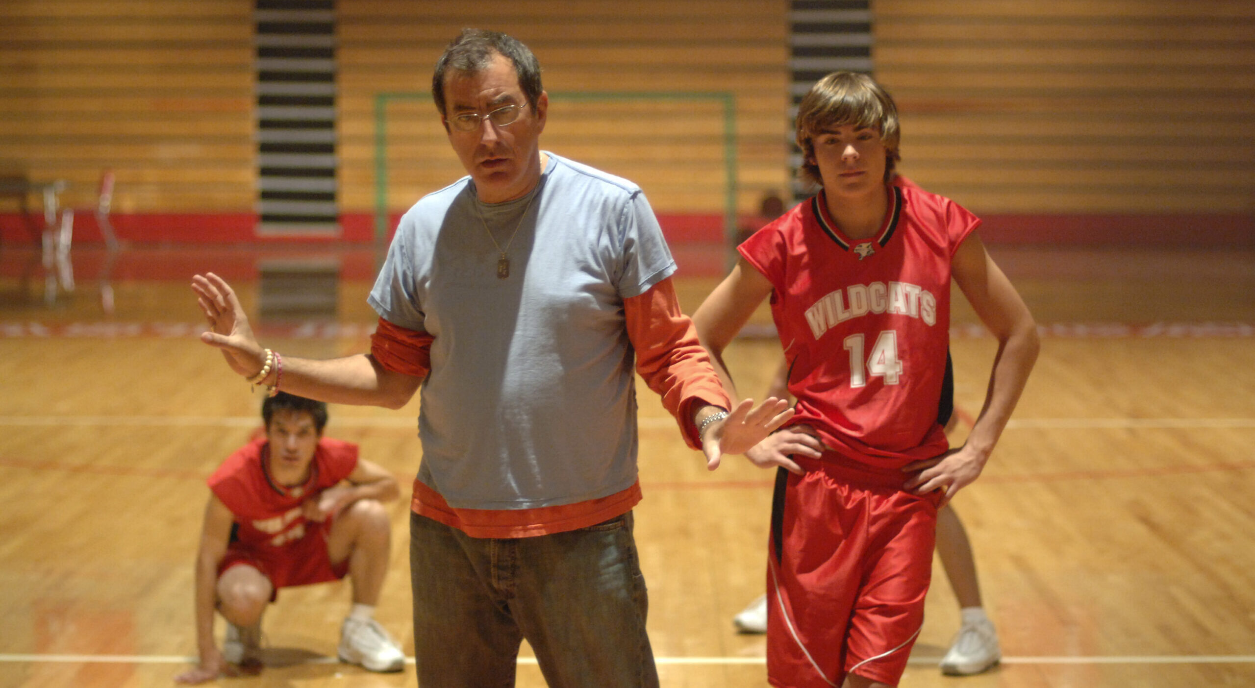 In a high school basketball gym, Kenny Ortega stands with his arms gently posed at either side. He looks slightly down and speaks to three dancers, clad in red and white basketball uniforms, who stand or crouch behind him.