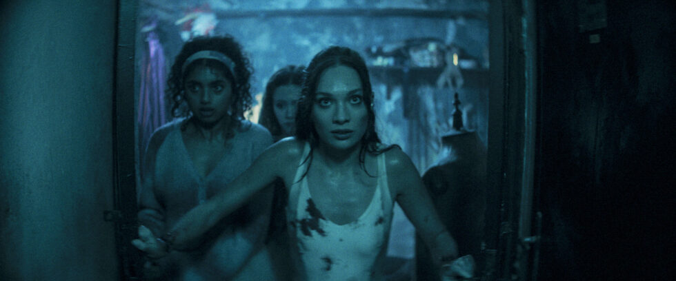 Three teenaged ballerinas, in bloodstained white costumes, burst through a doorway in a dark, dingy basement. The lighting is dark and blue-tinted.