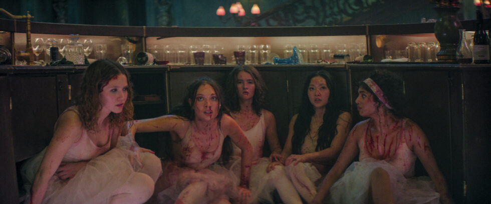 Five teenage ballerinas, wearing bloodstained white Romantic tutus, crouch behind a bar with alarmed expressions.
