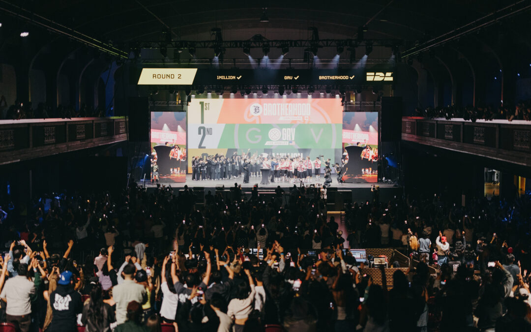 In a large, dark arena, a crowd cheers a group of dancers on a raised stage.