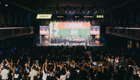 In a large, dark arena, a crowd cheers a group of dancers on a raised stage.