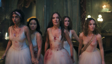 Five teenage ballerinas, wearing bloodstained white Romantic tutus, stand together in a dingy inn lobby with alarmed, but focused, expressions.