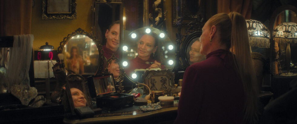 A woman with a sleek high ponytail paints her face white at a vanity with several mirrors. She looks at a young dancer's reflection in one of the mirrors and speaks to her, smiling menacingly.