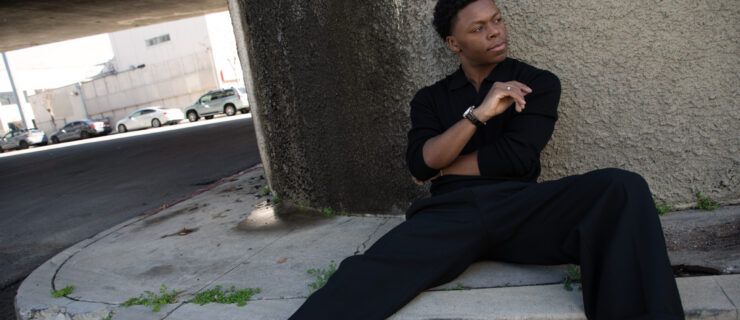 Rhyan Hill, in black slacks and a button-down rolled up at the elbow, sits casually on the ground of a parking garage. He leans back against a concrete wall, legs extended long in front of him with a slight bent, and folds his arms gently as he looks off to the side with a pensive expression.