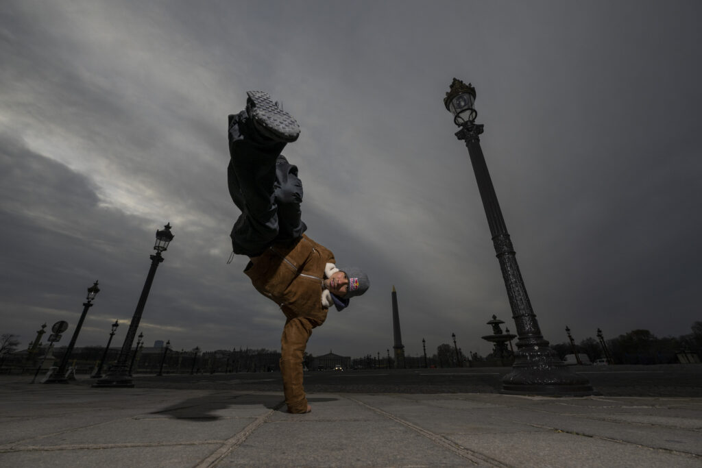 Victor Montalvo swings his legs up for a one-handed handstand. He dances in Paris' Place de la Concorde and wears a gray beanie, a brown jacket, black pants, and black boots.