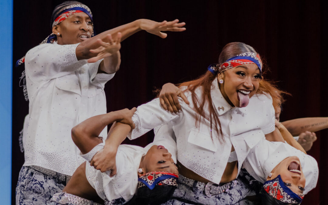 Maya Ramey sticks her tongue out while supporting two other dancers as they bend back and look towards the audience. The team wears patterned pants, white button down shirts, and bandanas.