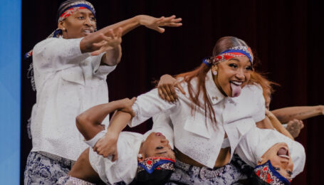 Maya Ramey sticks her tongue out while supporting two other dancers as they bend back and look towards the audience. The team wears patterned pants, white button down shirts, and bandanas.