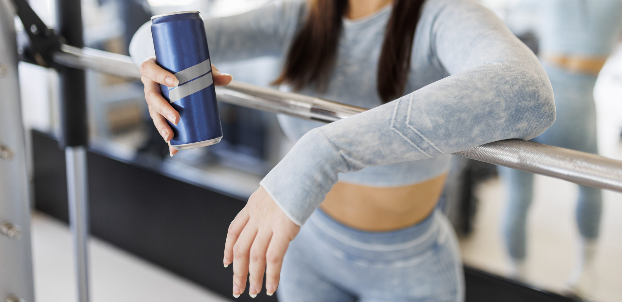 A woman rests her arms on a ballet barre in a gym, holding an energy drink in one hand.