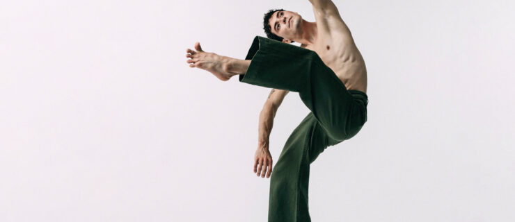 Jacob Beasley stands on one foot en relevé while lifting the other leg in attitude front and leaning laterally the same direction.