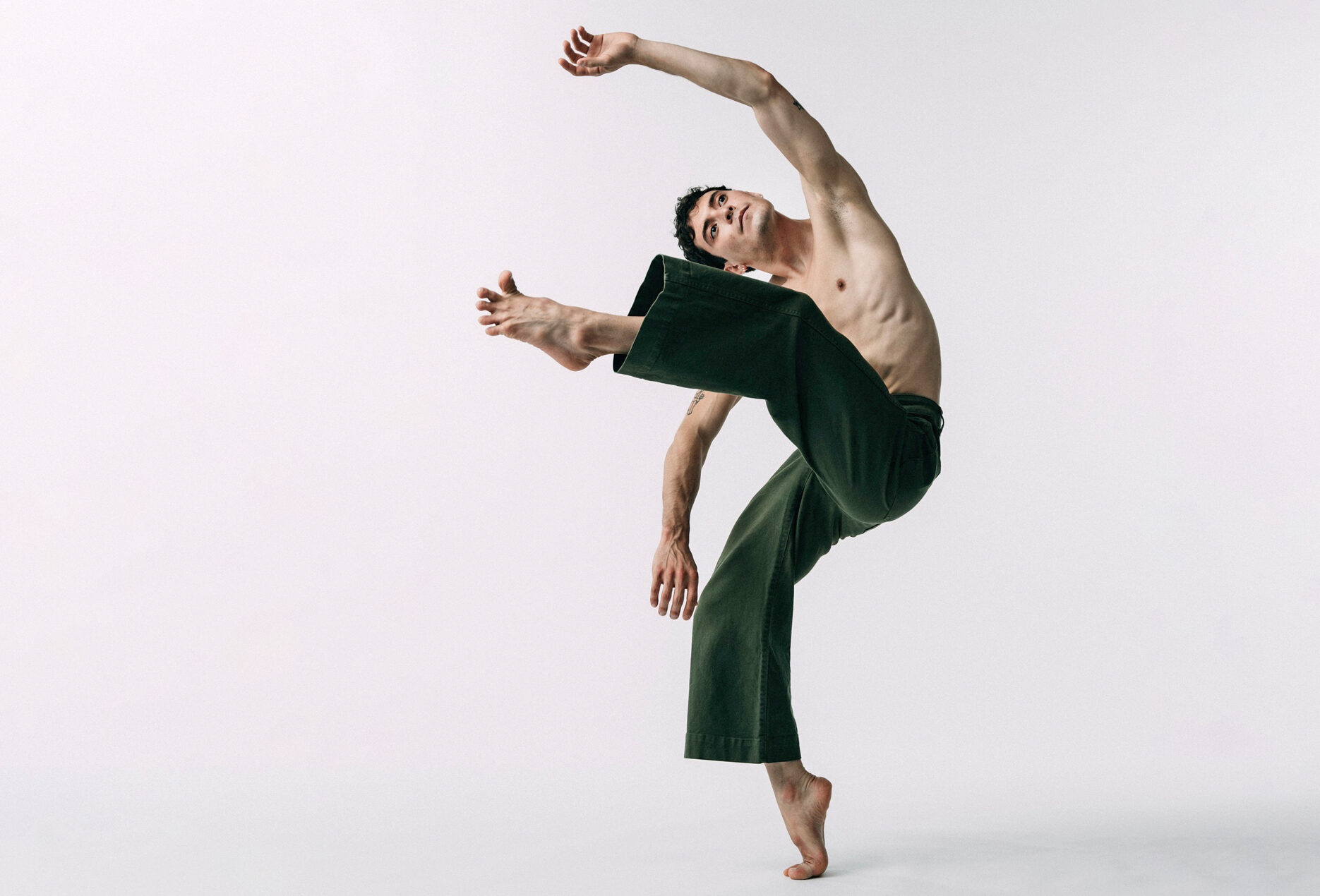 Jacob Beasley stands on one foot en relevé while lifting the other leg in attitude front and leaning laterally the same direction.