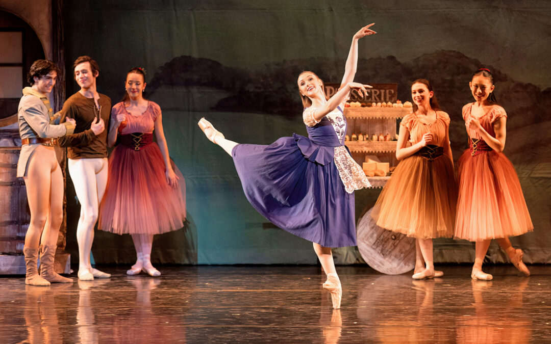 Audrey Lynn performs an attitude derrière in a peasant style dress. Fellow dancers stand behind her as characters in the scene.