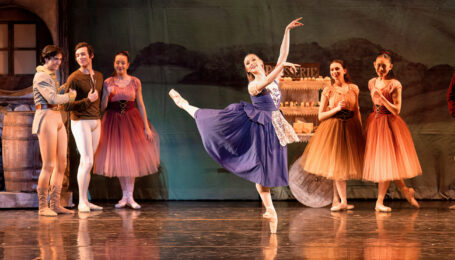Audrey Lynn performs an attitude derrière in a peasant style dress. Fellow dancers stand behind her as characters in the scene.