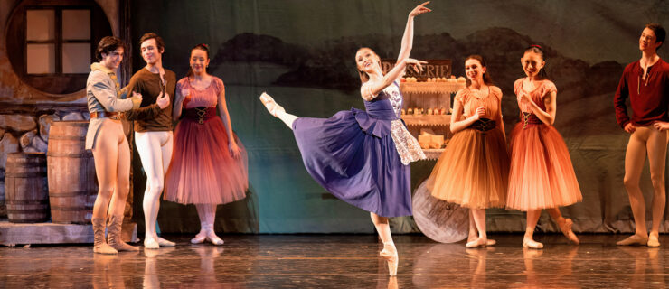 Audrey Lynn performs an attitude derrière in a peasant style dress. Fellow dancers stand behind her as characters in the scene.