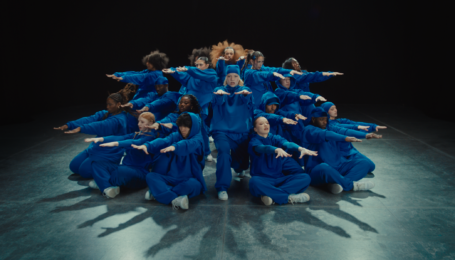 A large group of dancers pose in a circle, facing outward. The bottom row of dancers crouch and extend their arms out straight in front, palms down. The other levels do the same with slightly bent or straight legs. They wear bright blue matching sweatsuits.