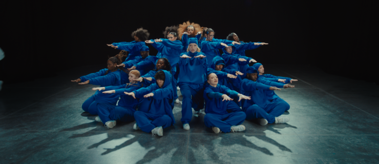 A large group of dancers pose in a circle, facing outward. The bottom row of dancers crouch and extend their arms out straight in front, palms down. The other levels do the same with slightly bent or straight legs. They wear bright blue matching sweatsuits.