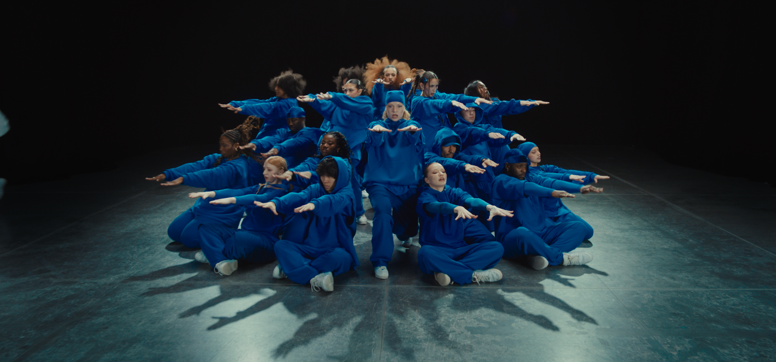 A large group of dancers pose in a circle, facing outward. The bottom row of dancers crouch and extend their arms out straight in front, palms down. The other levels do the same with slightly bent or straight legs. They wear bright blue matching sweatsuits.
