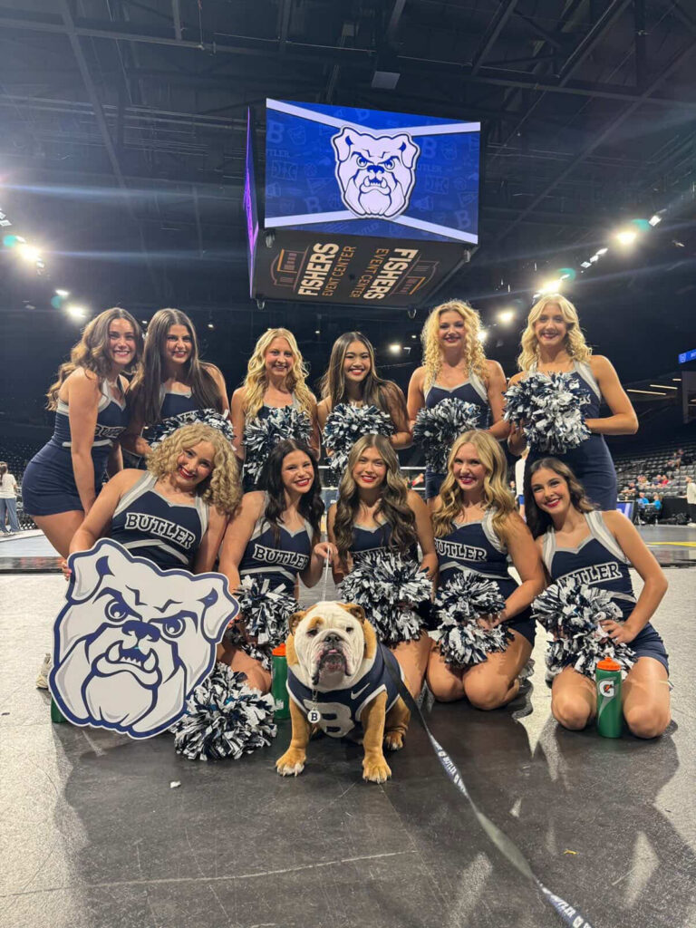 Megan Dang and her fellow Butler University Dance Team members pose for a group photo around the Butler bulldog mascot. They are in a sports facility and hold pom-pons.
